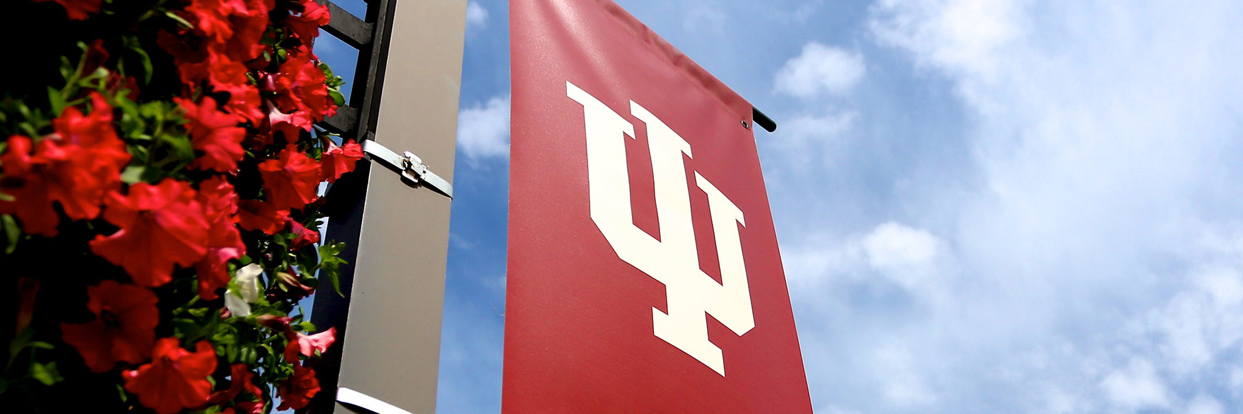 A red, IU trident banner hangs on a lamp post with a bright blue sky behind it.